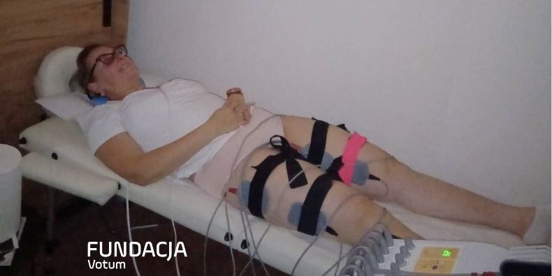 Woman lying on a medical bed with leg straps and electrodes attached, undergoing therapy; control device nearby (Fundacja Votum logo).