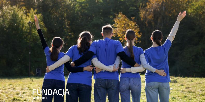 Five people with blue shirts have arms around each other in a sunny field, symbolizing teamwork and unity.
