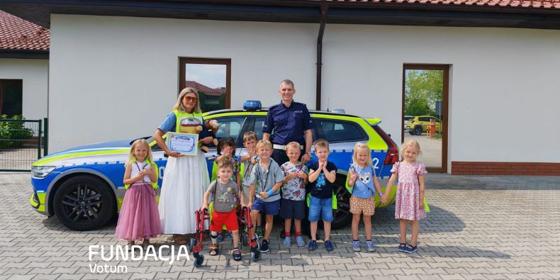 Group of children and two adults (a police officer and a woman) posing in front of a blue-yellow police car; one child holds a certificate of achievement.