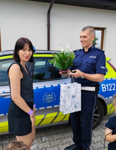 Police officer in a blue uniform hands a bouquet of white lilies and a gift bag to a smiling woman beside a police car; children watch.