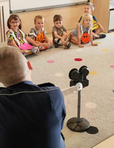 Five preschoolers sit in a semicircle on a colorful classroom carpet, wearing bright costumes, as an adult operates a camera on a stand nearby.