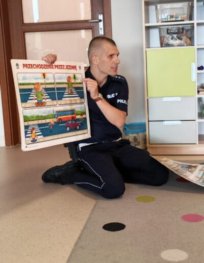 Police officer in uniform sitting on the floor, presenting a large illustrated road-safety poster about crossing the street to children