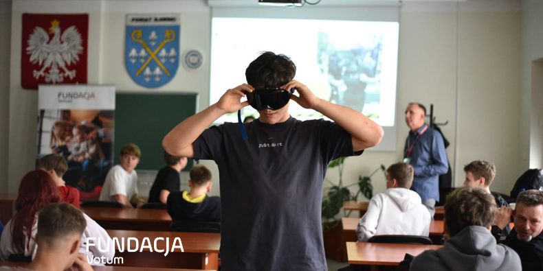 Young man in a classroom puts on a virtual reality headset while others watch. Projector screen with a blurred image is in the background.